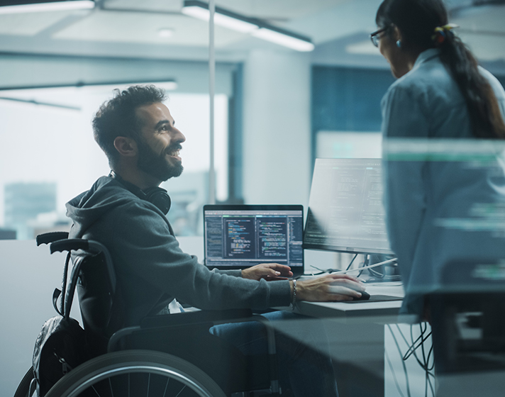Disability-Friendly Office: Brilliant IT Programmer with Disability in a Wheelchair Talks with Colleague while Working on Computer. Software Engineers Develop Innovative App, Program.